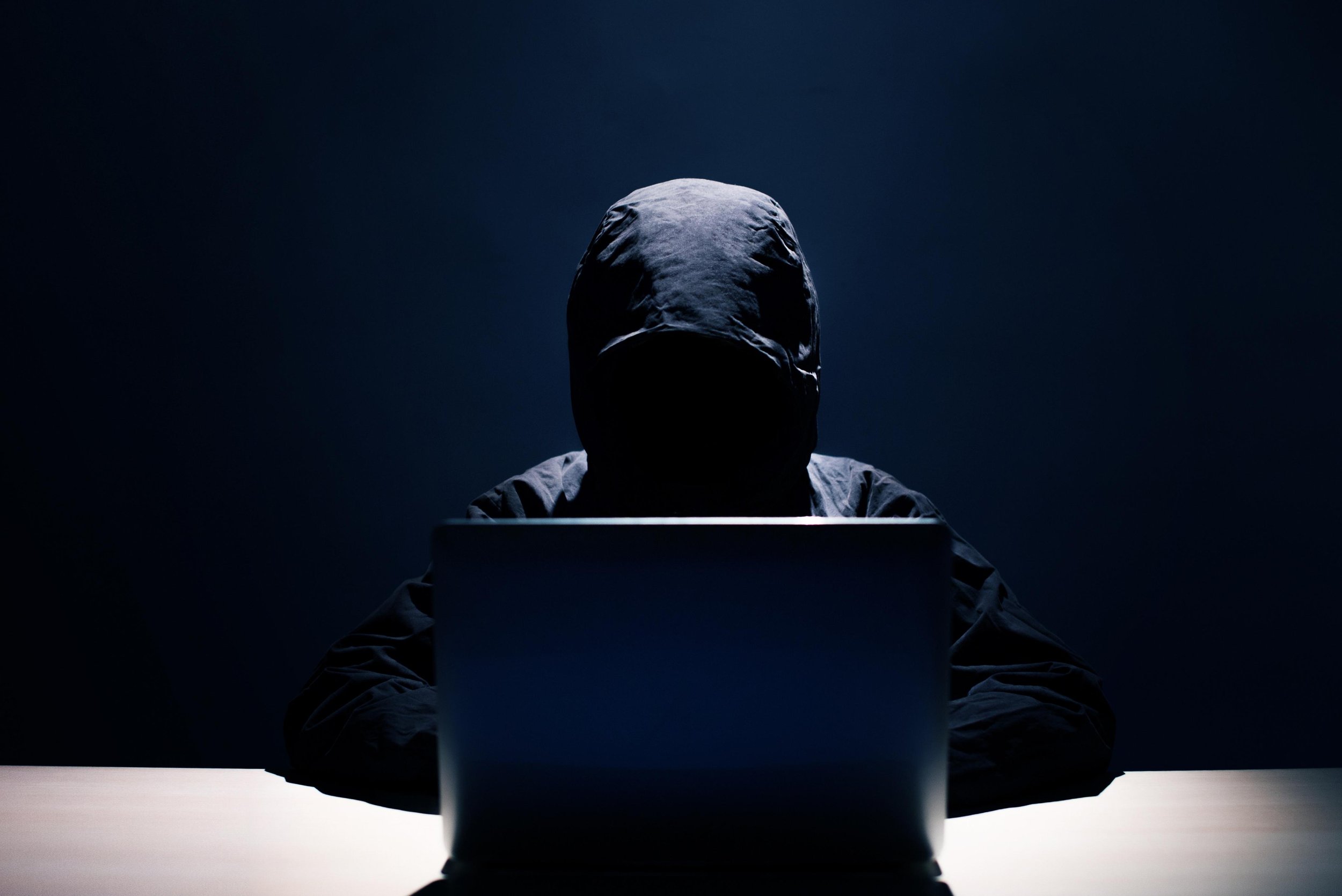 A lone hooded figure silhouetted at a computer in a darkened, blue-lit room